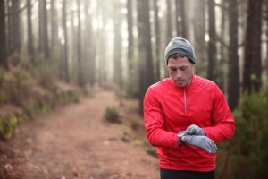 Trail runner man looking at heart rate monitor watch running in cold forest wearing hat and gloves. Male jogger running training in woods.